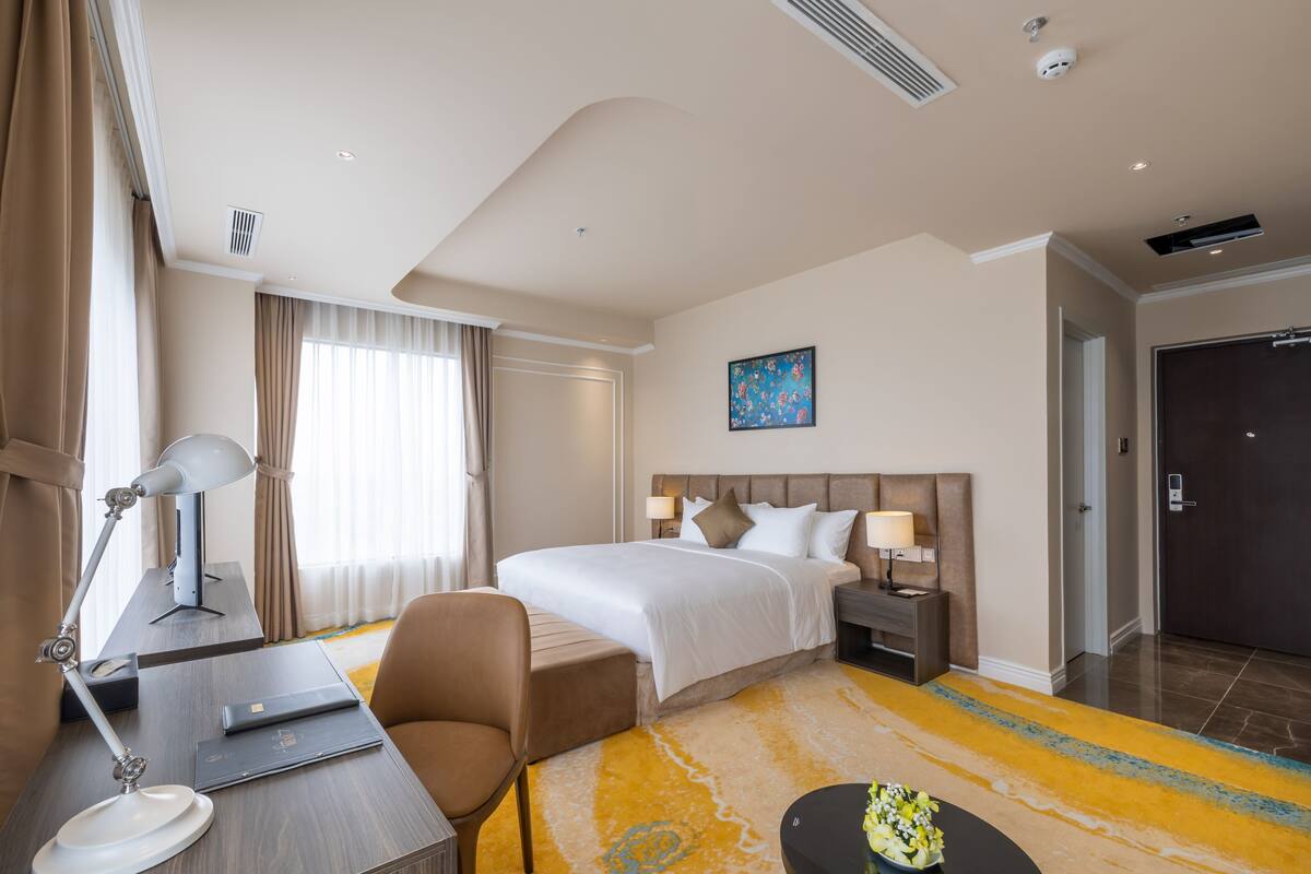 A hotel room with a bed, bedside tables, a desk with a lamp, a chair, and a small table with a vase.