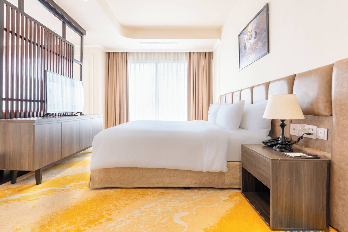 A hotel room with a large bed, a bedside table with a lamp, a TV on a stand, and a wooden wardrobe.