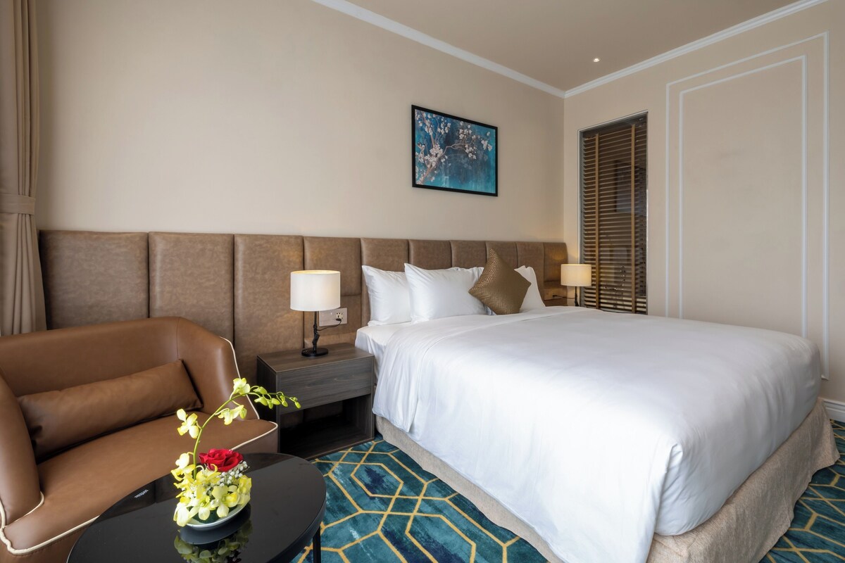 A hotel room with a large bed, a brown leather sofa, a small table with a vase of flowers, and a nightstand with a lamp.