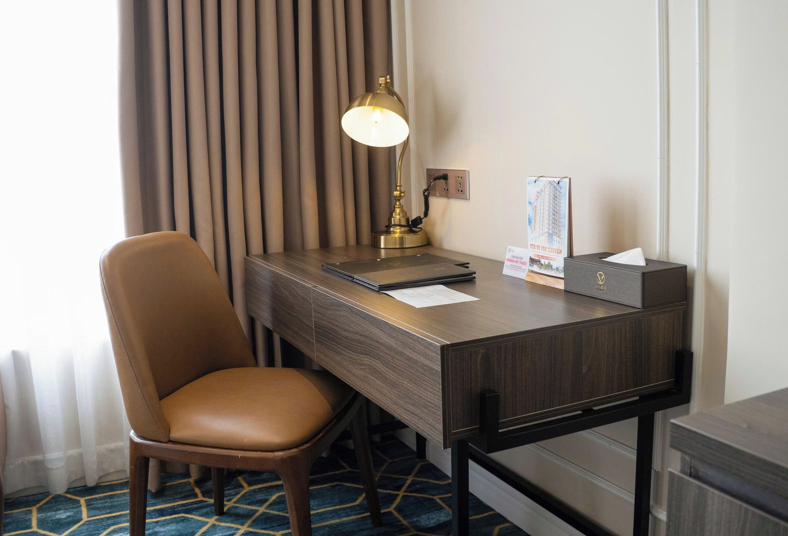 A hotel room with a desk, chair, lamp, and a small table with a tissue box and a brochure.