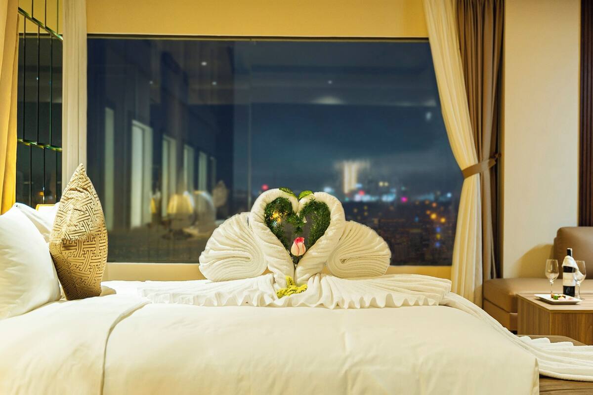 A hotel room with a bed featuring heart-shaped towels and a view of a cityscape at night.