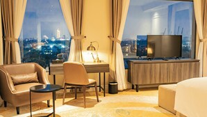 A hotel room with a large window, a desk with a lamp, a television, a sofa, and a chair. - La Vela Hue Hotel (Hue)