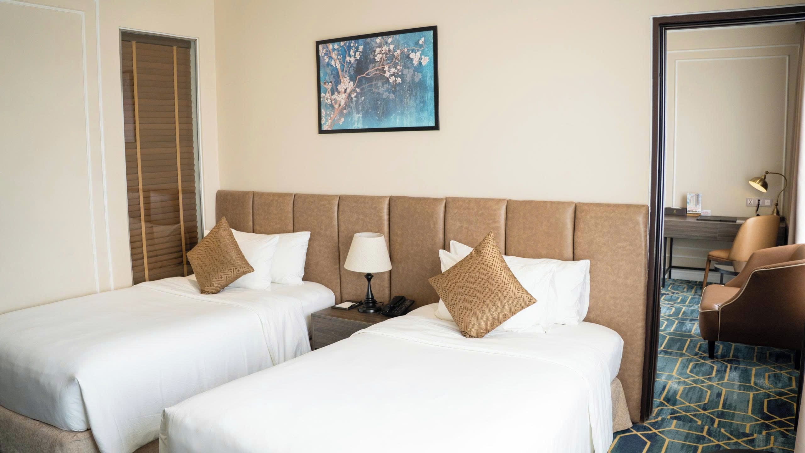 Ein Hotelzimmer mit zwei Betten, einem Nachttisch, einer Lampe, einem Gemälde und einem Schreibtisch mit Stuhl.