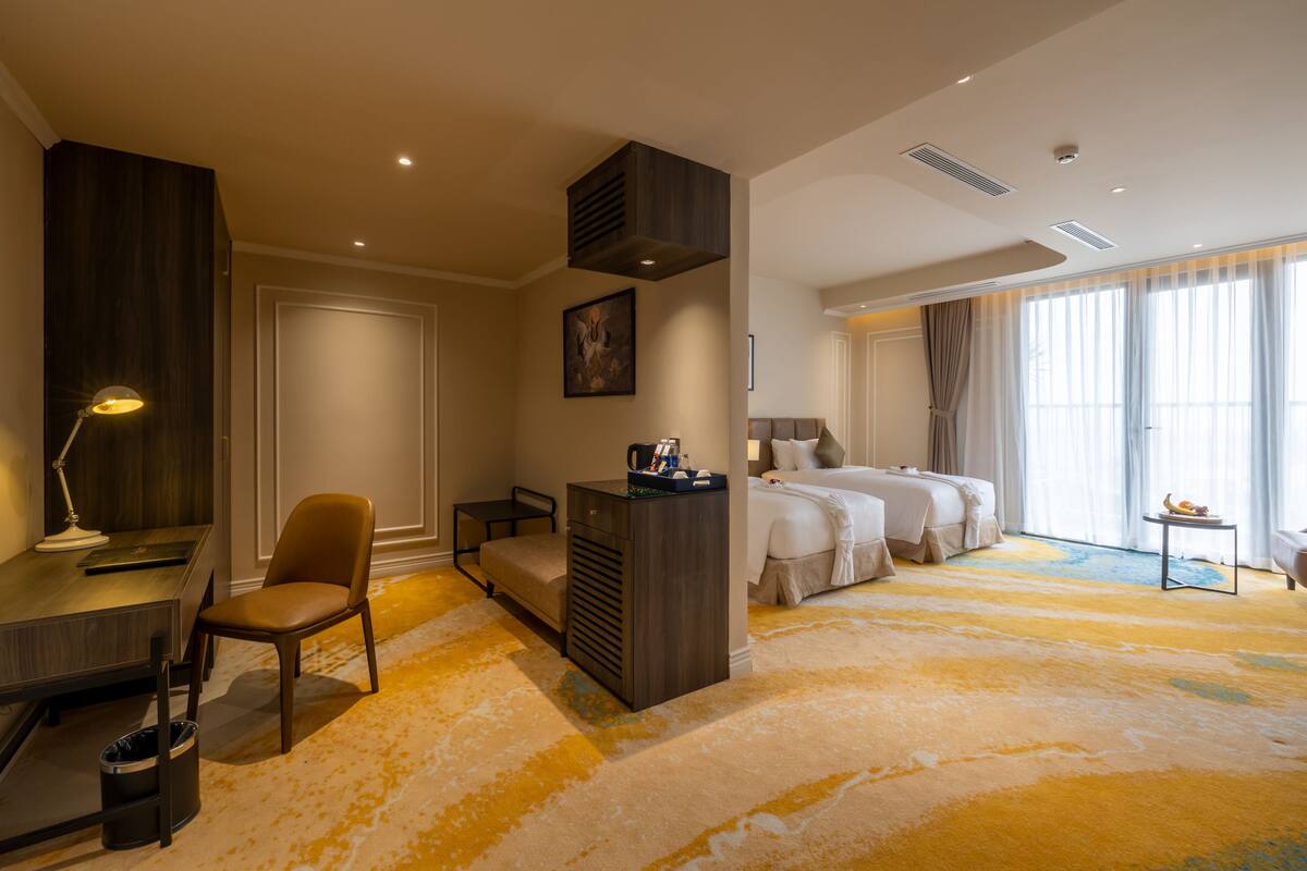 A hotel room with a desk, chair, two beds, a small table, and a large window with curtains.