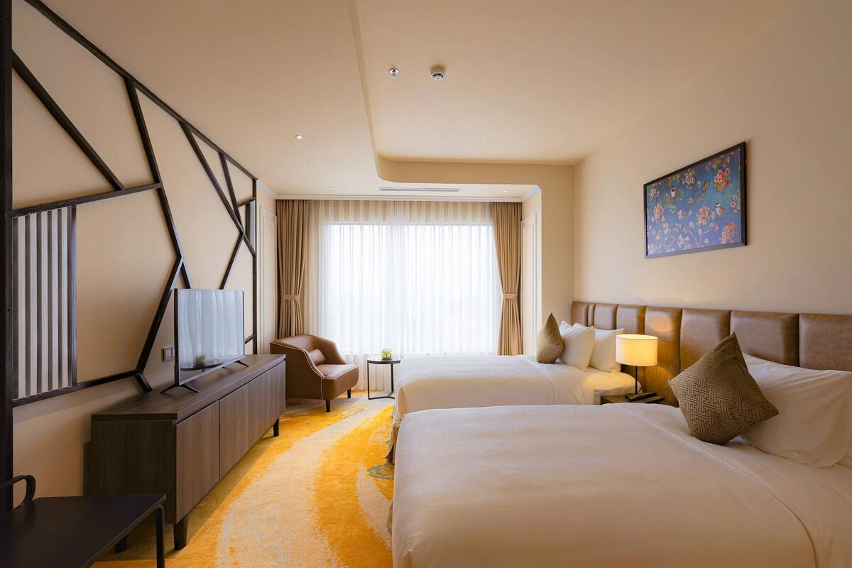A hotel room with two beds, a desk with a flat-screen TV, a chair, a small table, and a painting on the wall.