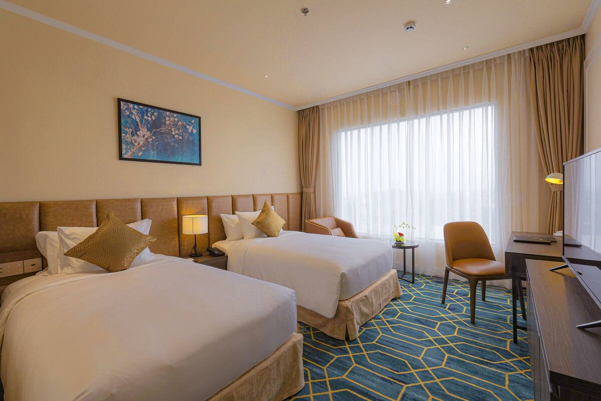 A hotel room with two beds, a desk, a chair, a lamp, and a painting on the wall.