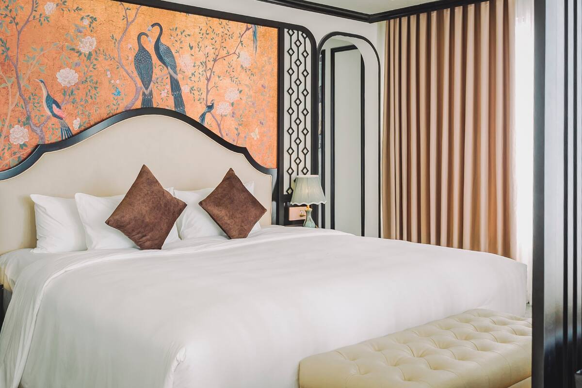A neatly made bed with white and brown pillows, a tufted bench, and a wall with a decorative painting of birds.