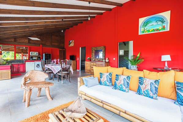 A spacious living area with a red wall, a sofa with tropical pillows, a dining table with chairs, and a kitchen area with red cabinets.