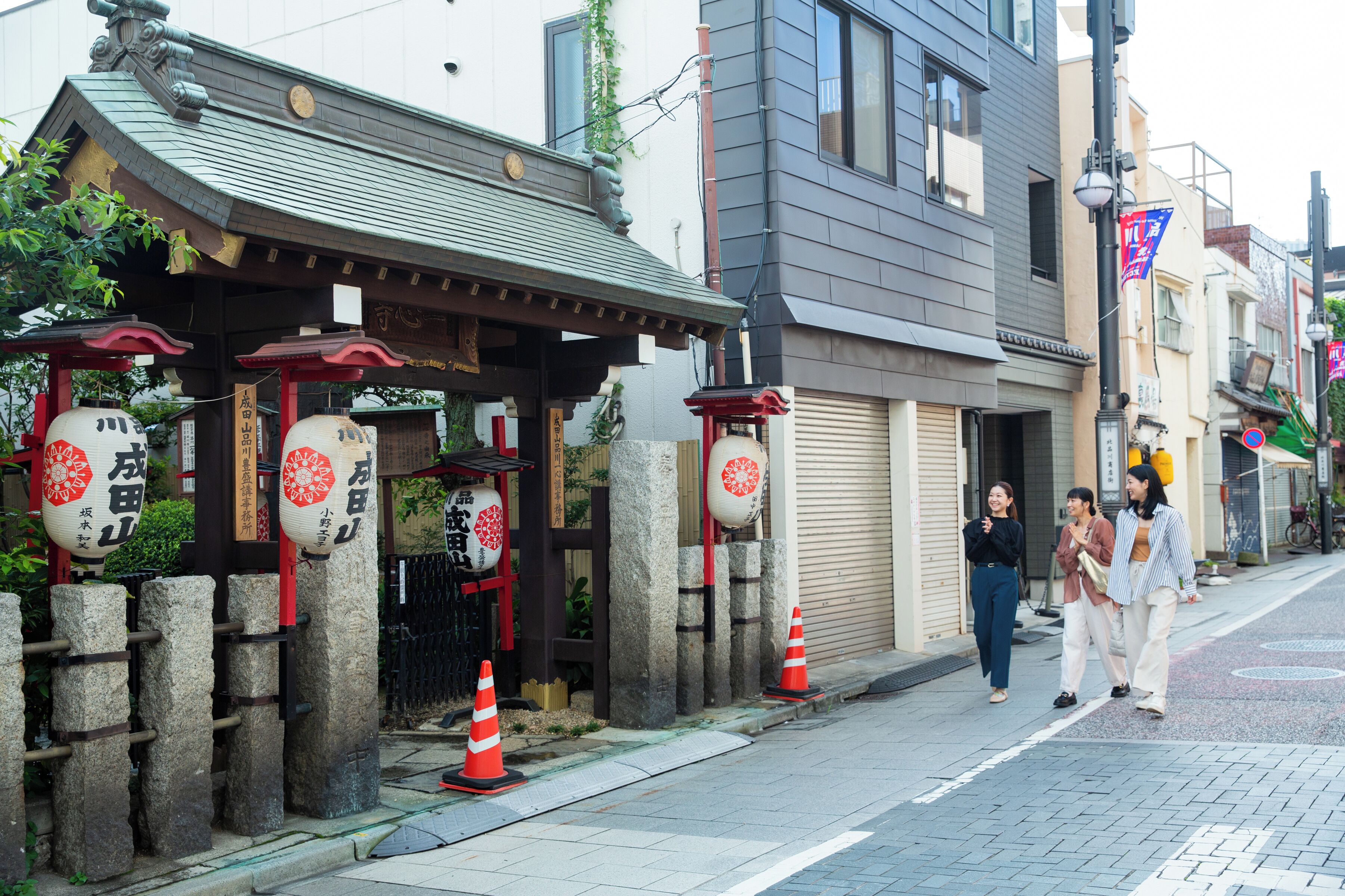 A traditional Japanese building with stone pillars and red lanterns, a sidewalk, and pedestrians.