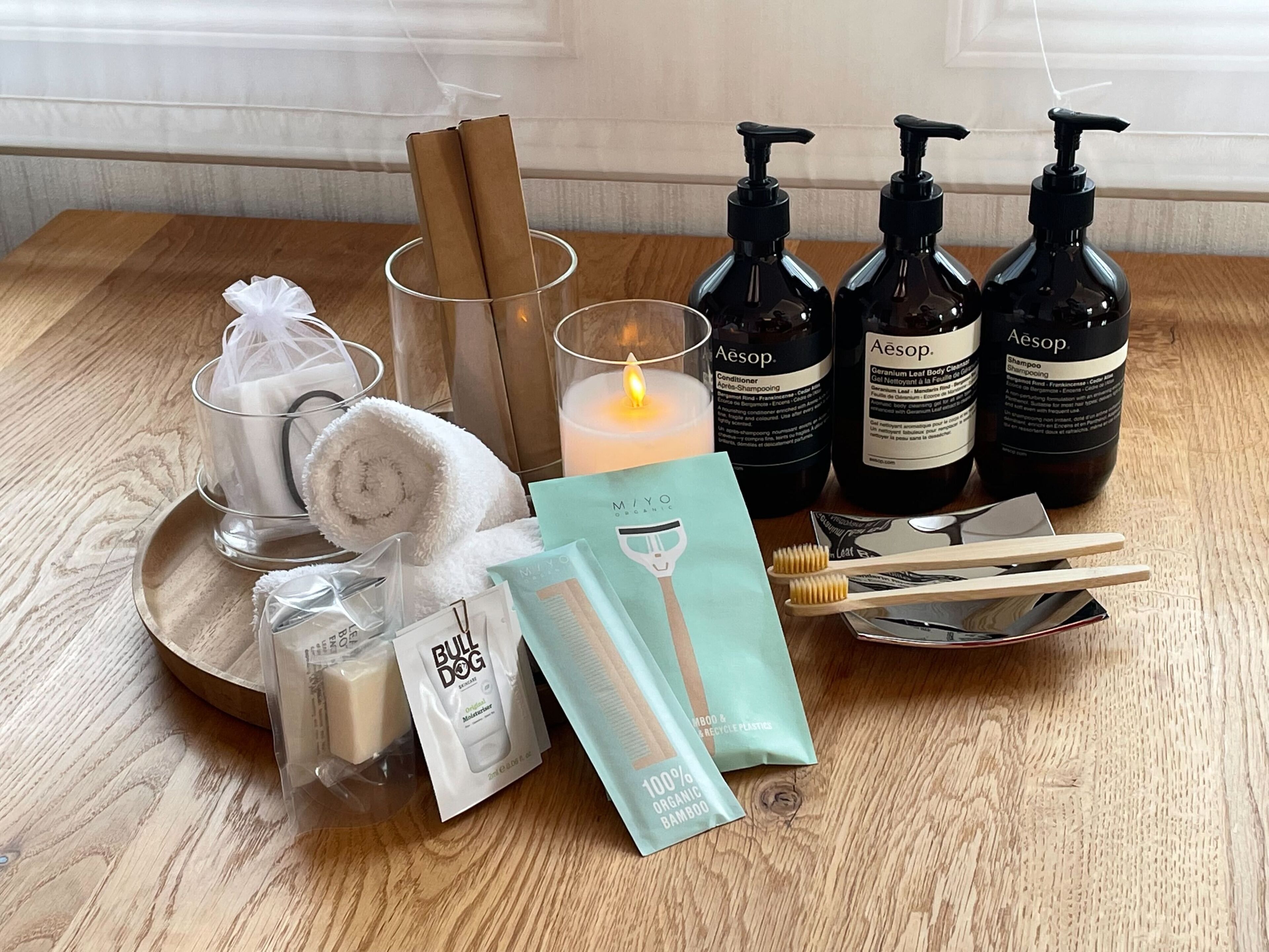 A wooden table with toiletries including a candle, bottles of shampoo and conditioner, a towel, and a toothbrush.