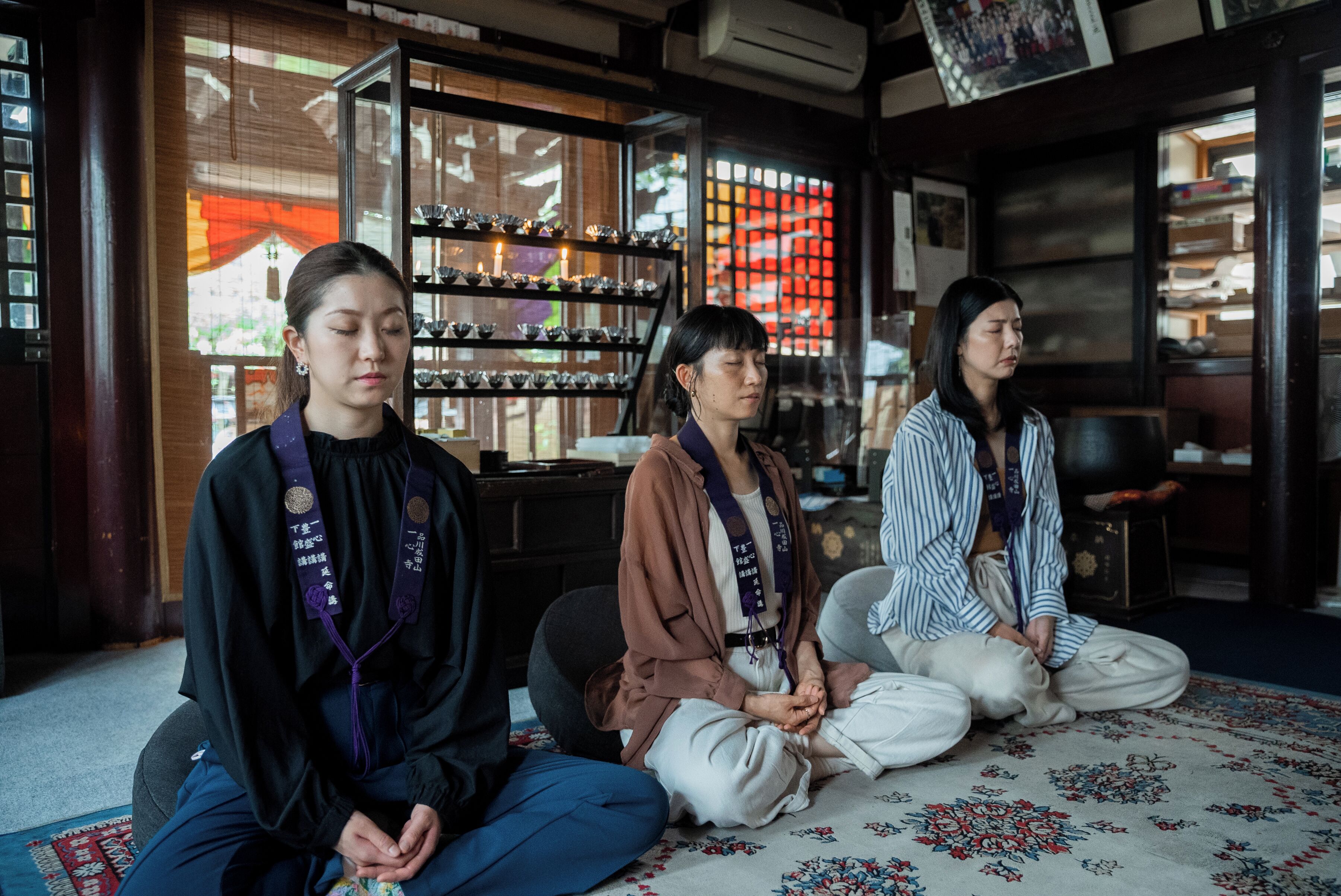 Three individuals seated on a patterned rug in a room with wooden furniture and shelves with glassware.