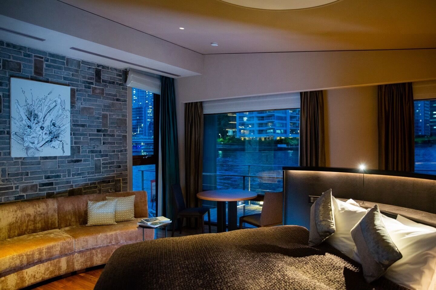 A modern hotel room with a brick accent wall, a large bed, a sofa, a small table, and a balcony with a view of the city at night.