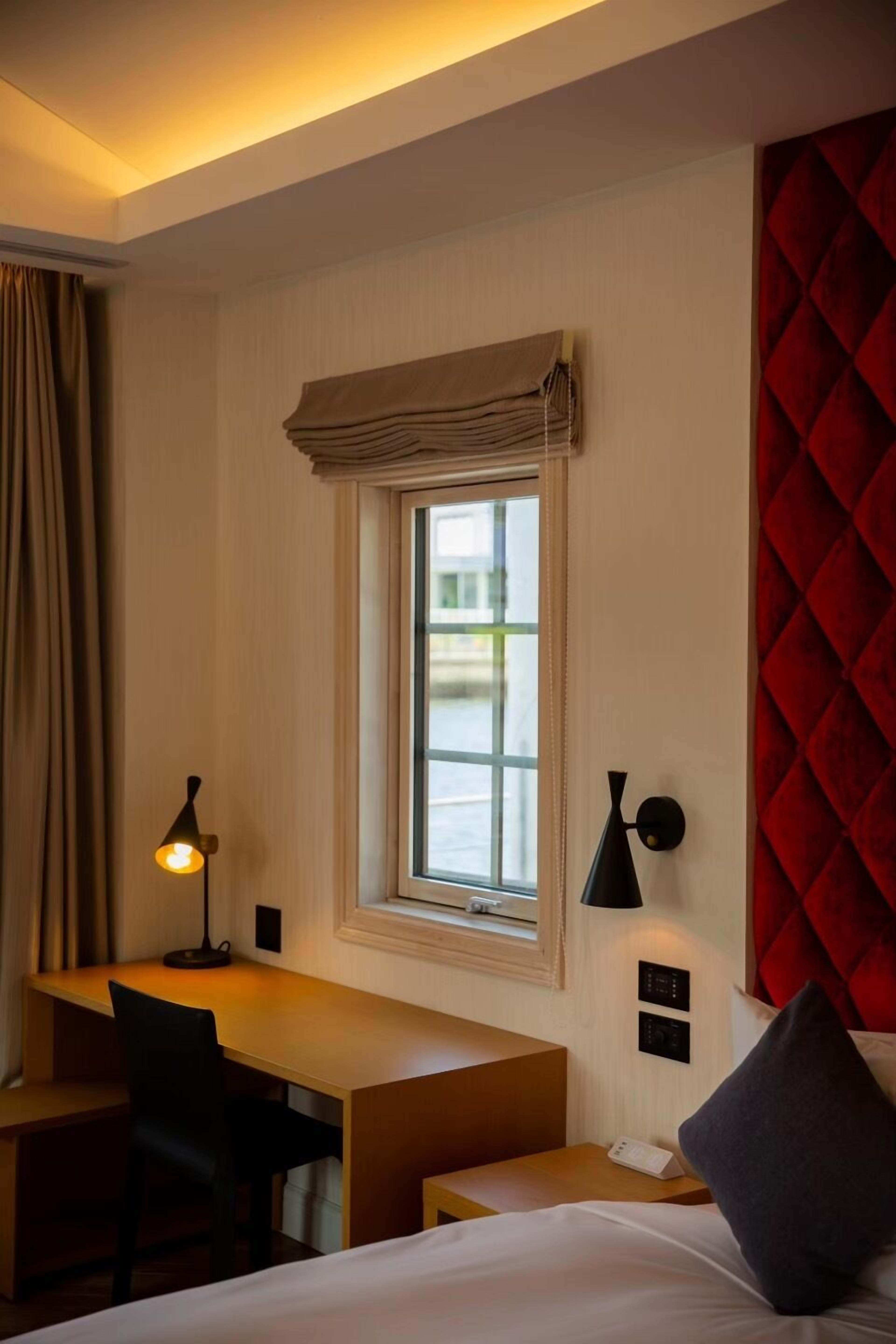 A hotel room with a bed, a desk, a chair, a window with curtains, a red accent wall, and a wall-mounted lamp.