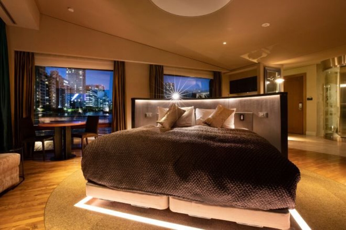 Una moderna camera d'hotel con un ampio letto, vista sulla città, zona pranzo e ascensore in vetro.