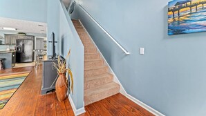 A modern kitchen with a refrigerator, stove, and bar stools. A staircase leads to a living area with a painting of a sunset over water. - Fun and Sun Vacation Townhome (Panama City Beach)