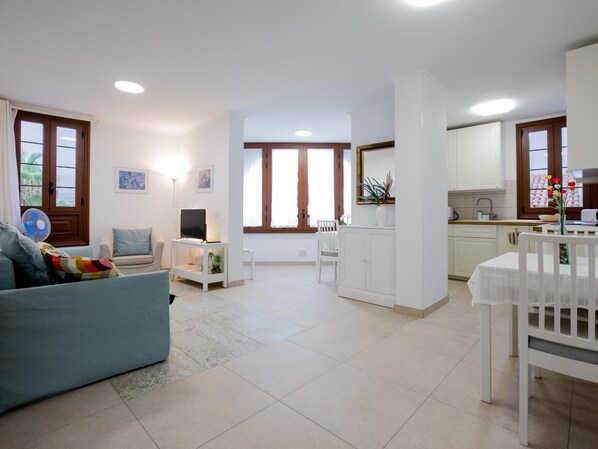 A modern living room with a sofa, TV, and fan, leading to a kitchen with a dining table and chairs.