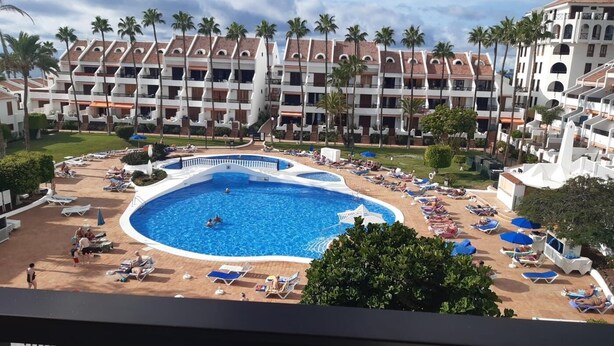 A hotel with a large central pool surrounded by sun loungers and palm trees.