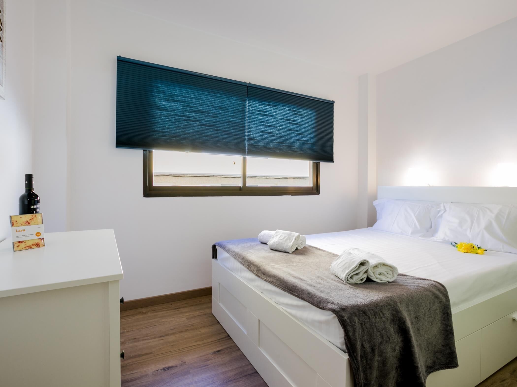 A bedroom with a bed, a window with blinds, a bedside table, and a bottle of wine.