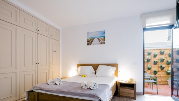 Une chambre à coucher avec un lit en bois, du linge de lit blanc, une table de chevet, un tableau accroché au mur et un balcon agrémenté de plantes en pot.