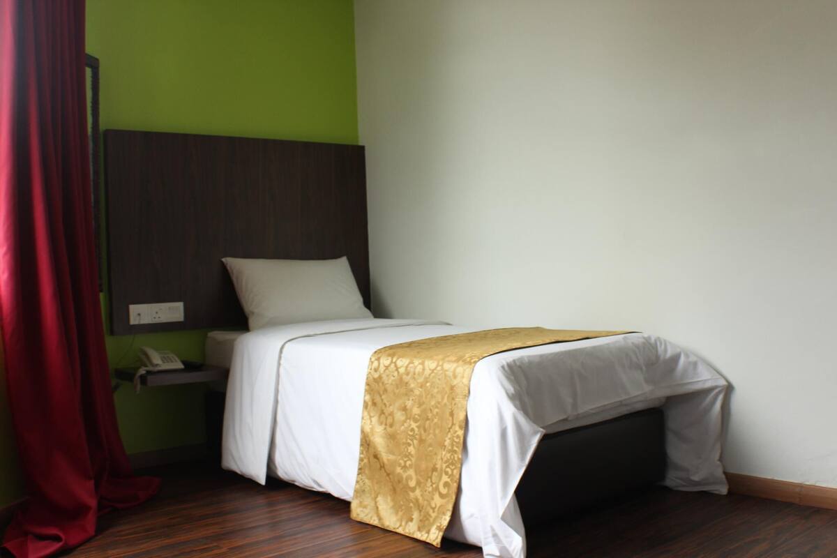 A single bed with a white mattress and a gold throw blanket, a bedside table with a phone, and a red curtain.