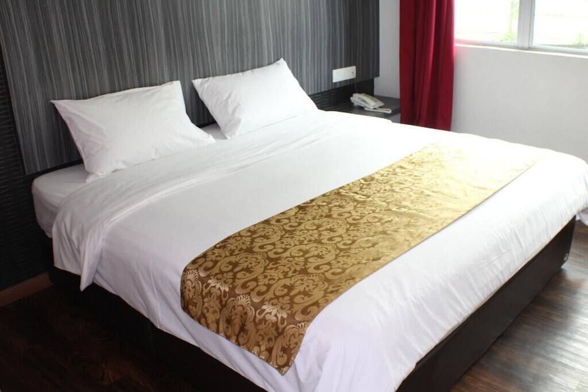 A neatly made bed with white linens and a gold-patterned throw, a bedside telephone, and a window with red curtains.