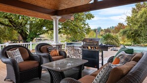 Eine Terrassengruppe mit Korbmöbeln, einem Stein-Barbecue und Blick auf ein Schwimmbad.