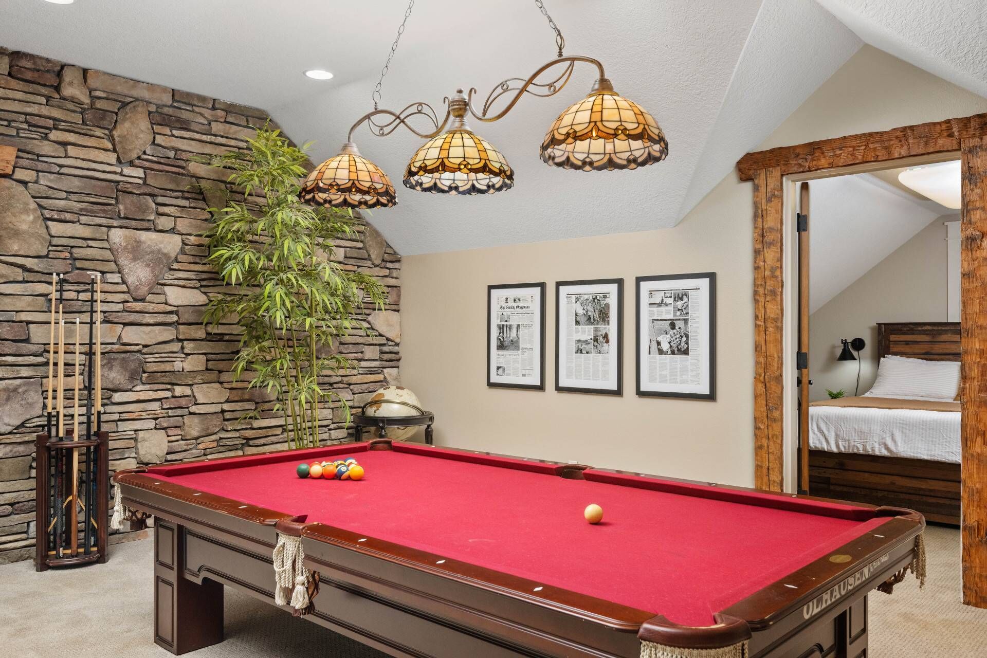 A pool table with a red felt surface, a stone wall, and three framed newspaper clippings on the wall.
