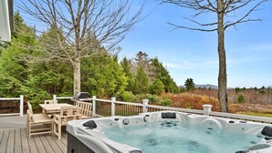 A hot tub on a wooden deck with a table and chairs, surrounded by trees and a view of the landscape.
