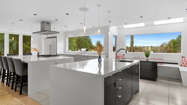A modern kitchen with a large island, bar stools, and a view of the outdoors through large windows.