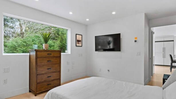 Un dormitorio moderno con una cama grande, un aparador, un televisor montado en la pared y una ventana con vistas a zonas verdes.