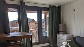 A modern living room with a wooden table, a sofa, a lamp, and a view of the city through a large window with curtains. - LES Glaciers Studio 25m2 (Les Belleville)