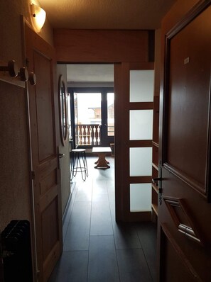 Un pasillo con una puerta de madera a la derecha, una mesita con sillas y vistas al exterior a través de una ventana.