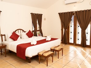 A hotel room with a large bed, two bedside tables, a wooden bench, and a window with curtains. - Jasminn Villas South Goa (Betalbatim)