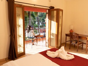 A hotel room with a bed, a desk, a chair, and a balcony with a view of greenery and buildings. - Jasminn Villas South Goa (Betalbatim)