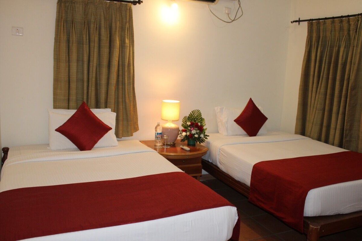 A hotel room with two single beds, each with a red pillow and a red blanket. There is a nightstand with a lamp, a small table with a vase of flowers, and a wall socket.