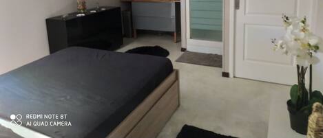 Une chambre à coucher moderne, avec un lit, une commode, une coiffeuse et une salle de bain visible à travers une porte en verre.