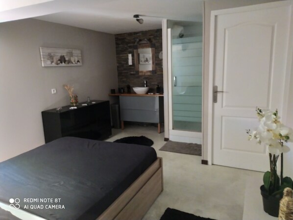 A modern bedroom with a bed, a dresser, a vanity, and a bathroom visible through a glass door. - Appartement Quartier Calme (Perpignan)