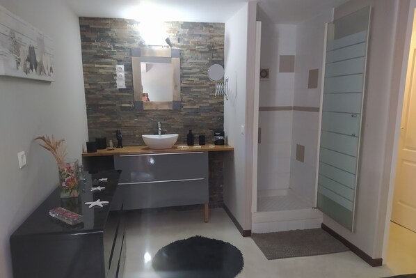 A modern bathroom with a stone backsplash, a large mirror, a sink, and a shower area. - Appartement Quartier Calme (Perpignan)