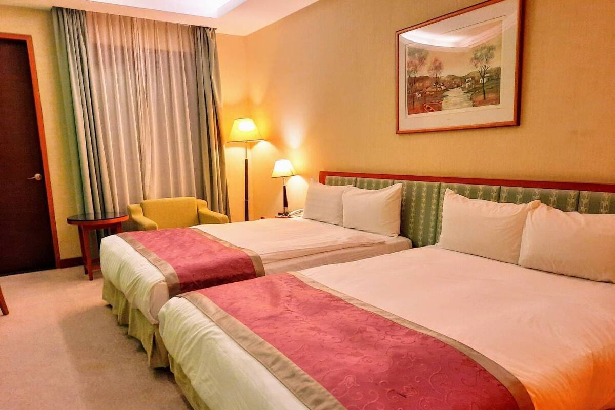 A hotel room with two beds, a chair, a lamp, and a painting on the wall.