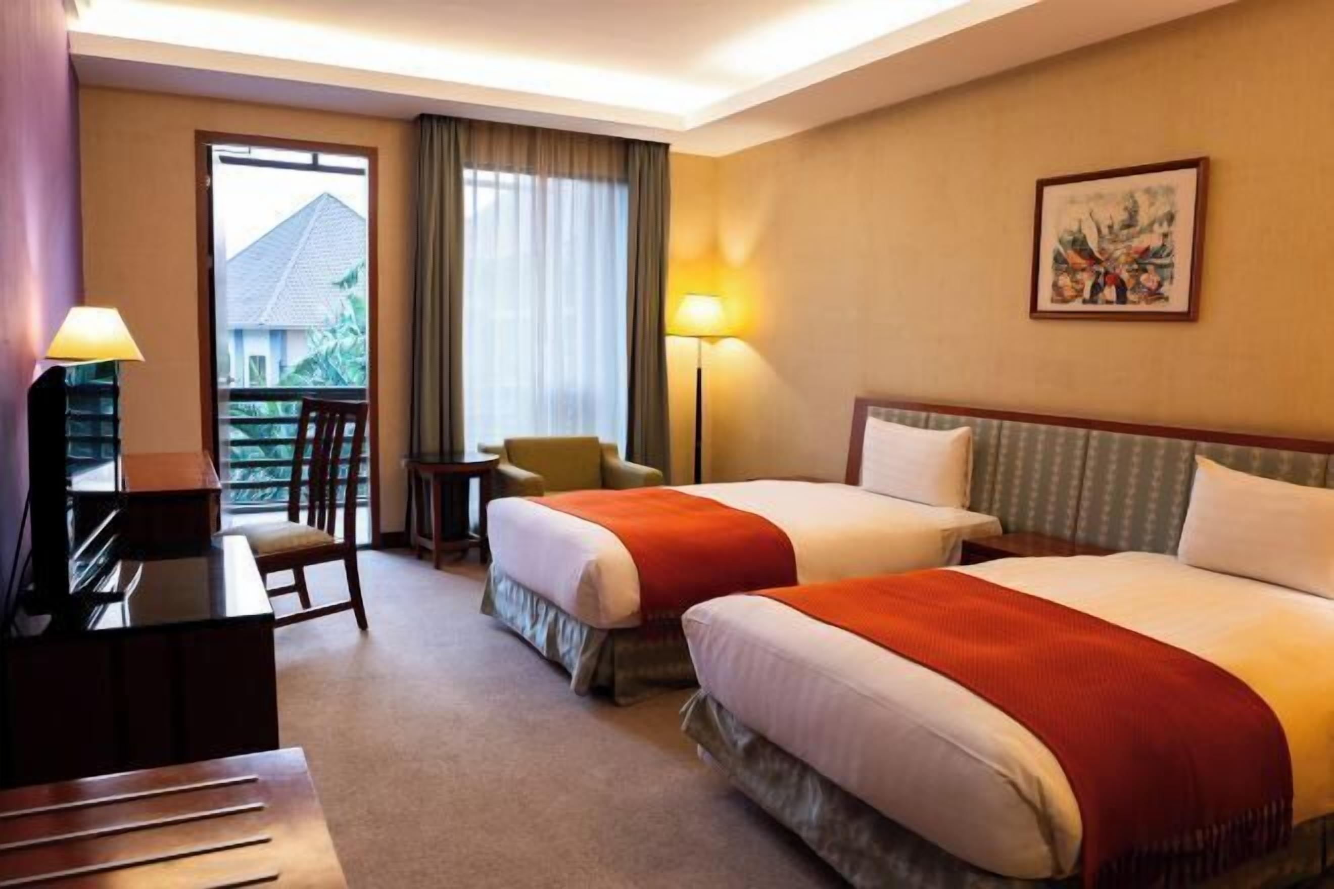 A hotel room with two beds, a desk, a chair, a lamp, and a painting on the wall.