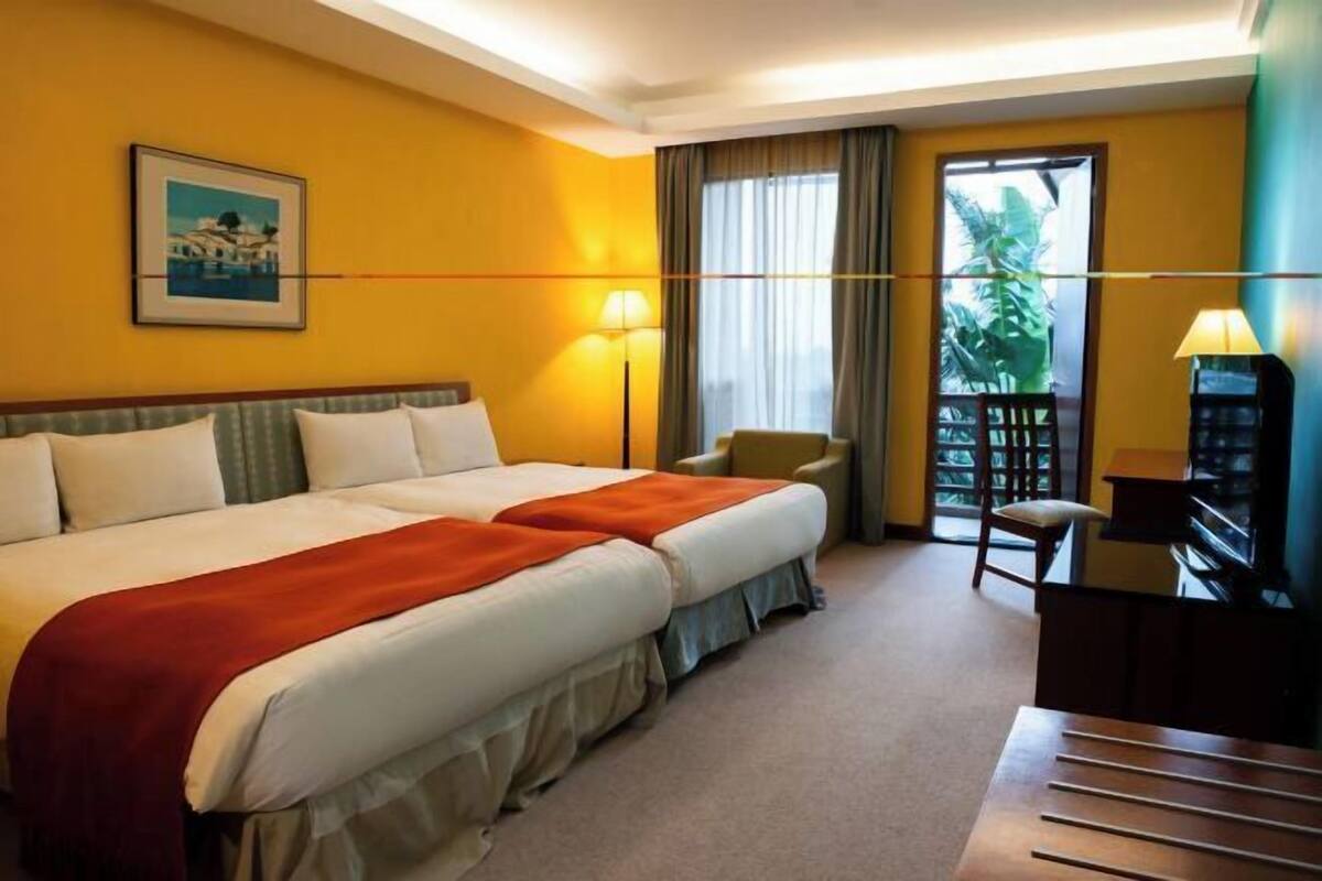 A hotel room with two beds, a desk, a chair, a lamp, and a window with curtains.