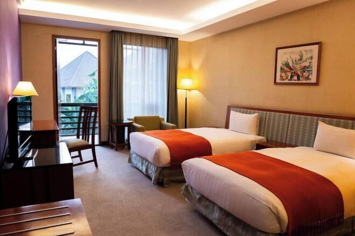 A hotel room with two beds, a desk, a chair, a lamp, and a painting on the wall.