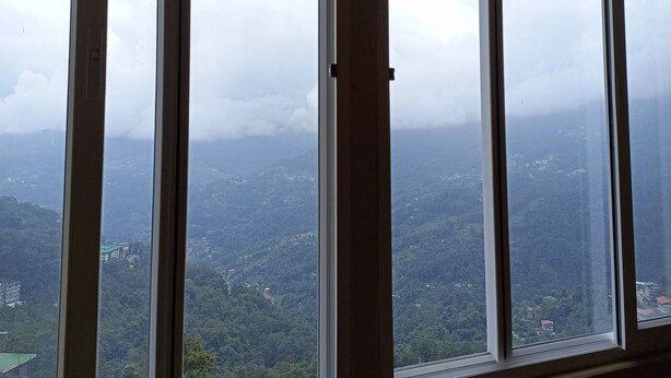 A view through a window with multiple panes, displaying a mountainous landscape with scattered buildings. - Mash Denzong Shiron (Gangtok)