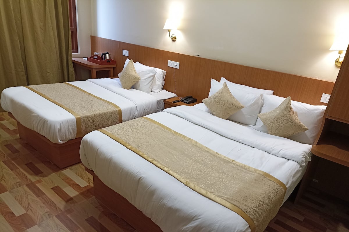 Quarto de hotel com duas camas, cada uma com colcha bege e almofadas brancas. Possui cabeceira de madeira, mesa de cabeceira com abajur e piso de madeira.