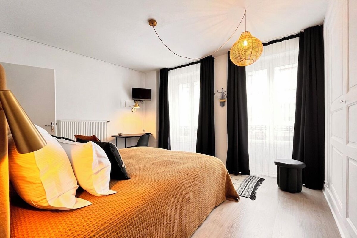 Une chambre d’hôtel avec un grand lit, un bureau, une chaise, un petit tabouret et une vue sur la ville par la fenêtre.