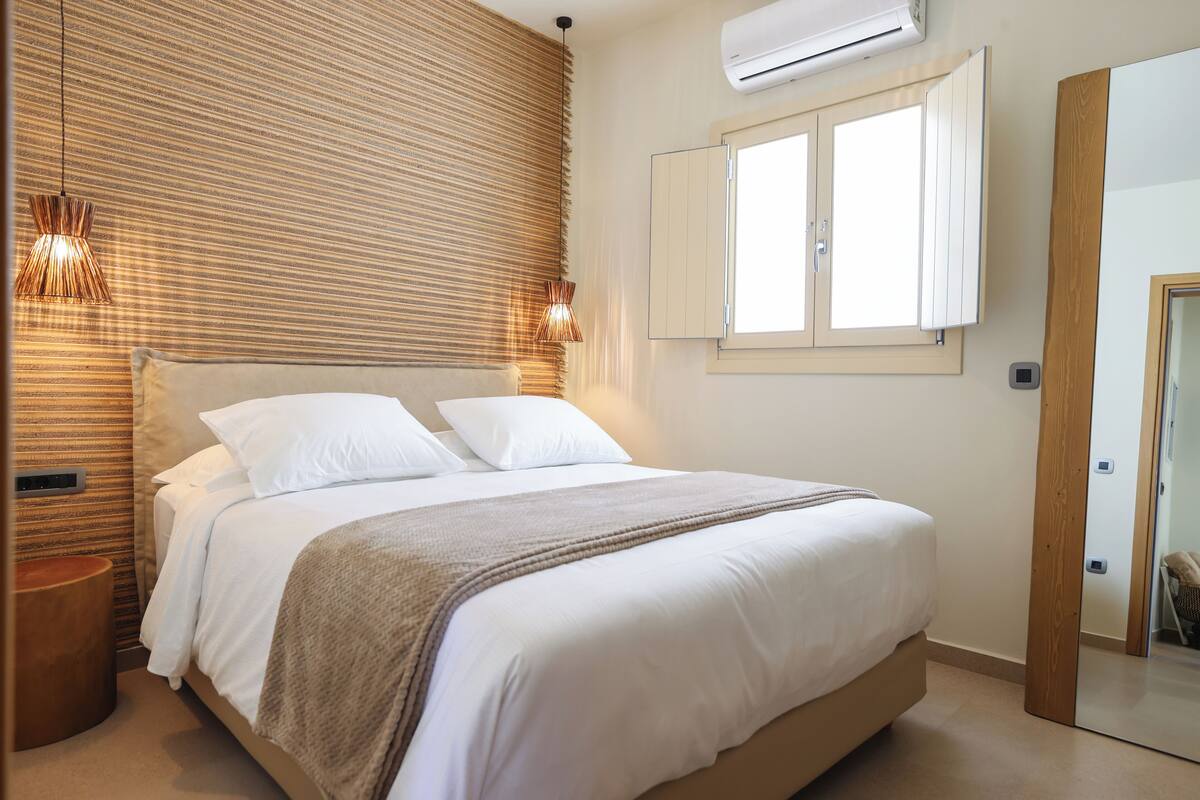 A neatly made bed with white linens and a beige throw blanket, a wooden slatted headboard, and a window with white shutters.