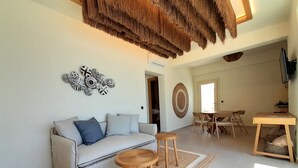 A living room with a sofa, coffee table, and a dining table with chairs. There is a decorative wall hanging and a woven ceiling. - LIFE SUITES (Mykonos)