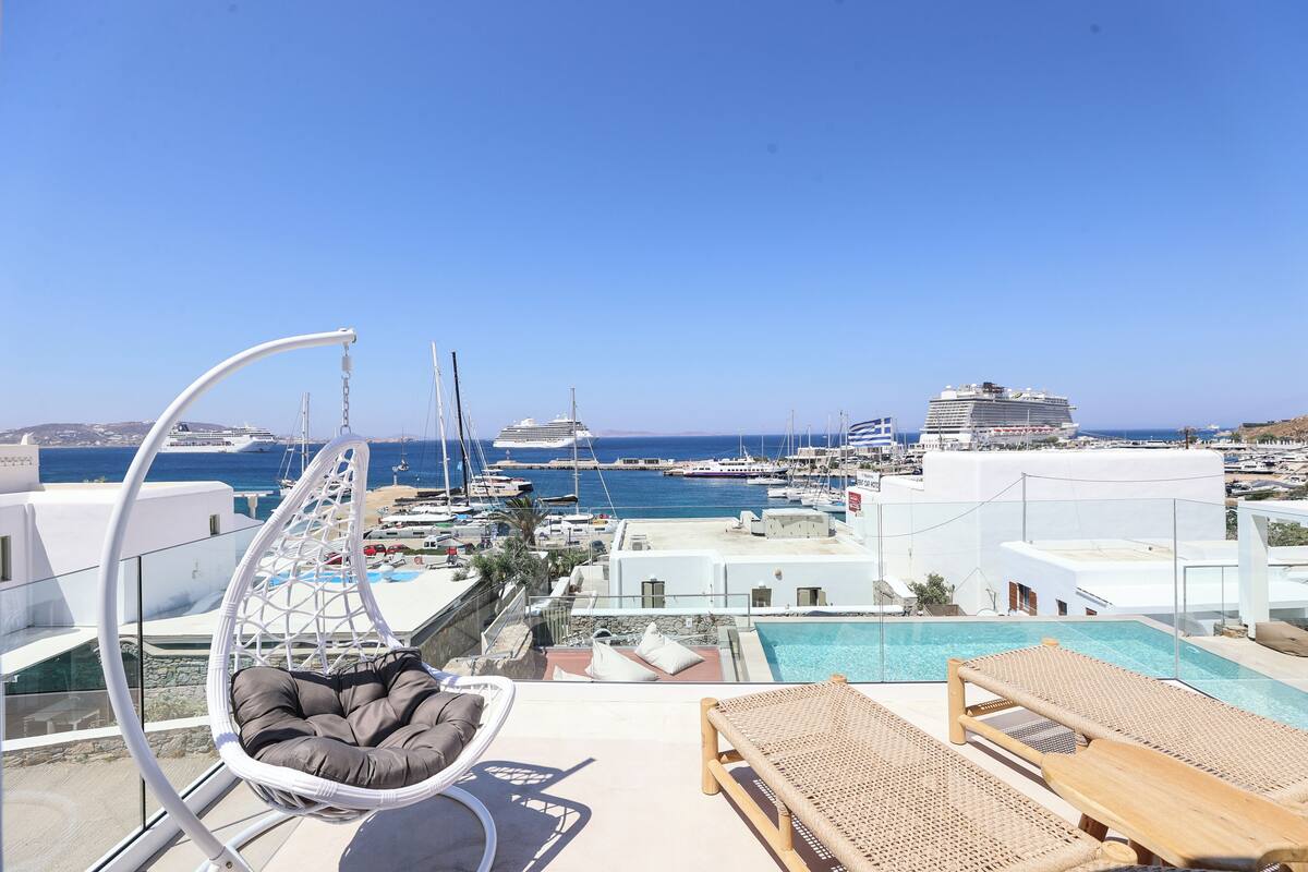 A rooftop pool area with a hanging chair and lounge chairs, overlooking a marina with boats and a cruise ship.