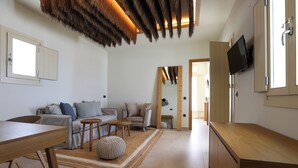 A modern living room with a sofa, coffee table, and ottoman, a wooden dining table, and a TV mounted on the wall. - LIFE SUITES (Mykonos)