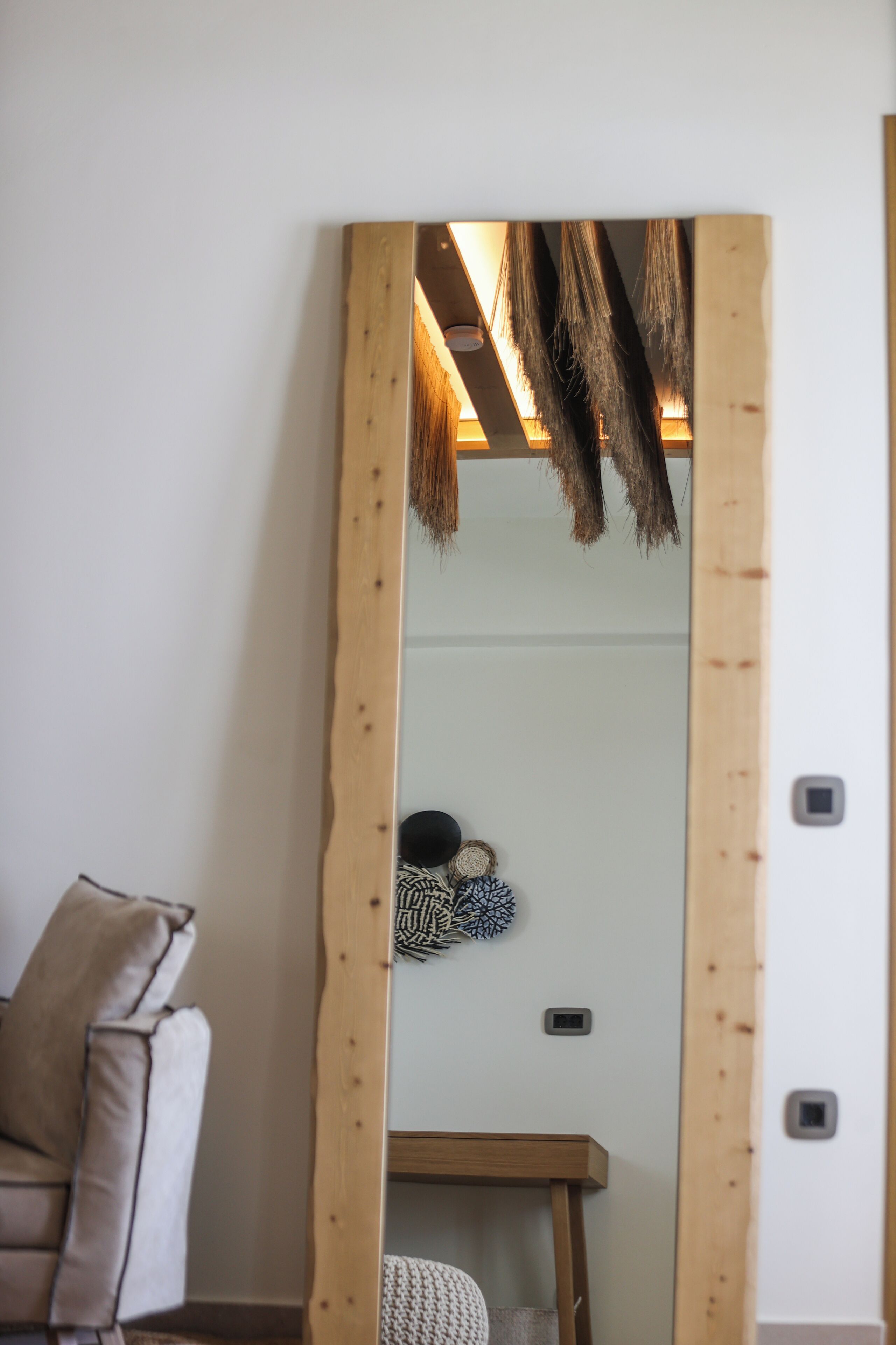 Een kamer met een grote spiegel die een plafond met houten balken en een hangende lamp weerspiegelt. In de weerspiegeling is een klein tafeltje met een gevlochten mand en decoratie te zien.